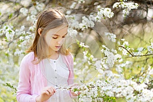 Adorable young girl having fun in blooming cherry garden on beautiful spring day. Kid hanging Easter eggs on blossoming cherry