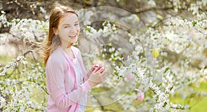 Adorable young girl having fun in blooming cherry garden on beautiful spring day. Kid hanging Easter eggs on blossoming cherry