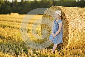 Adorable young girl having fun in a wheat field on a summer day. Child playing at hay bale field during harvest time. Kid enjoying