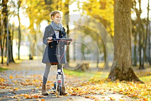 Adorable young girl riding her scooter in a city park on sunny autumn evening. Pretty preteen child riding a roller