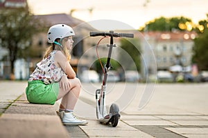 Adorable young girl riding her scooter in a city on sunny summer evening. Pretty preteen child riding a roller. Active leisure and