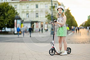 Adorable young girl riding her scooter in a city on sunny summer evening. Pretty preteen child riding a roller. Active leisure and