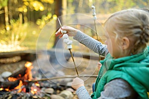 Adorable young girl roasting marshmallows on stick at bonfire. Child having fun at camp fire. Camping with children in fall forest