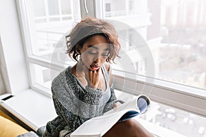 Adorable young girl with short curly hair absorbedly reading magazine, book sitting on windowsill, big white window. She