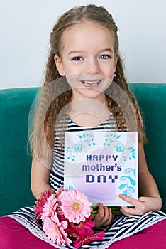 Adorable young girl sitting on a couch, holding bouquet of pink gerberas and Mother`s day card. Happy Mother`s Day