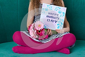 Adorable young girl sitting on a couch, holding bouquet of pink gerberas and Mother`s day card. Happy Mother`s Day