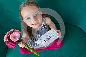 Adorable young girl sitting on a couch, looking up and holding bouquet of pink gerbera daisies and Mother`s day card.
