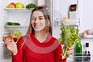 Adorable young woman with happy expression, dressed in red blouse, holds lettuce and sandwhich while stands at kitchen, near opene