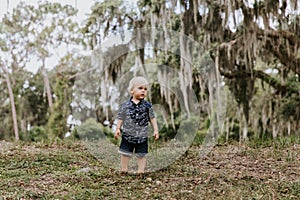 Adorably Happy and Cute Little Caucasian Toddler Baby Boy with Long Blond Hair Laughing, Playing, and Running Outside in Green Nat