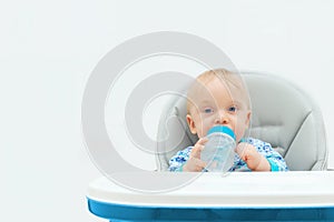 The adored child is sitting in a child`s chair, looking at the camera and holding a bottle of water