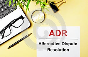 ADR Alternative Dispute Resolution is written in red on a white piece of paper on a light yellow background next to a laptop, pen