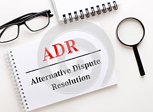 ADR Alternative Dispute Resolution is written in a white notebook on a light background near the notebook, black-framed glasses