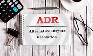 ADR Alternative Dispute Resolution is written in a white notepad near a calculator, coffee, glasses and a pen. Business concept