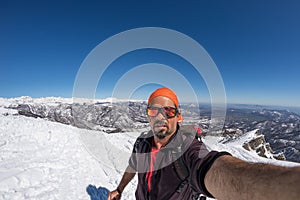 Adult alpin skier with beard, sunglasses and hat, taking selfie on snowy slope in the beautiful italian Alps with clear blue sky.