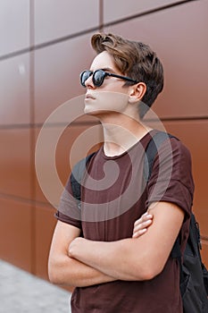 American young hipster man with backpack with trendy hairstyle in stylish sunglasses in fashionable brown t-shirt poses