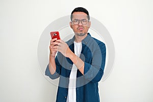 Adult Asian man looking at the camera with disappointed expression while using his phone