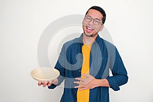 Adult Asian man showing empty bowl with happy expression while touching his stomach photo