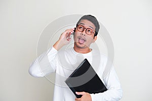 Adult Asian man showing funny ugly face while holding laptop and talking to someone on the phone