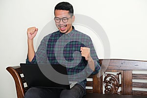 Adult Asian man sitting in a bench showing success gesture while looking to his laptop photo