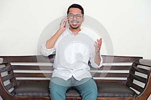 Adult Asian man sitting in a couch smiling while talking with someone in a phone call photo