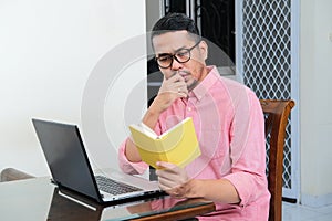 Adult Asian man sitting in front of his laptop while reading a book with serious expression photo