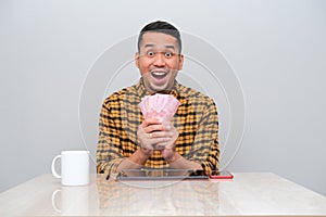 Adult Asian man sitting in working table with amazed expression while showing money that he hold