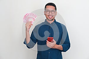 Adult Asian man smiling at the camera while holding phone and showing handful of money