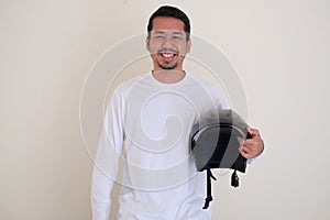 Adult Asian man smiling friendly while holding a motorbike helmet beside