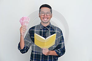 Adult Asian man smiling happy at camera while holding a book and money