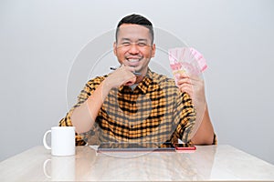 Adult Asian man smiling happy when sitting in his working table and showing money that he earn