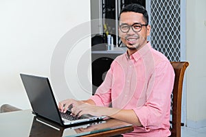 Adult Asian man smiling while working using laptop in his home photo