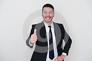 Adult Asian man wearing black suit and tie giving thumb up with happy expression