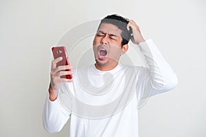 Adult Asian man yawning and scratching his head while holding his mobile phone photo