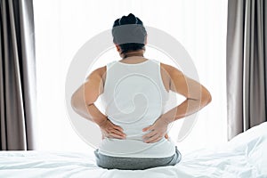 Adult Asian woman is sitting on the bed and holding her lower back suffering from injured back. Health care and back pain concept