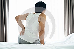 Adult Asian woman is sitting on the bed and holding her lower back suffering from injured back. Health care and back pain concept