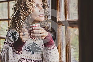 Adult attractive curly blonde woman at home enjoying winter holidays drinking from decorated mug and looking outside wht winwos photo