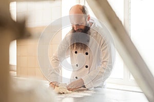 Adult baker with long beard in white uniform standing in his workplace and prepare the bread dough with hands kneading the dough