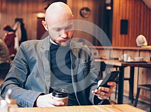 Adult bald gloomy man drinking coffee from paper cup and using mobile phone at cafe