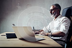 Adult bearded businessman wearing a classic glasses and working at the wood table in modern loft.Elegant man making