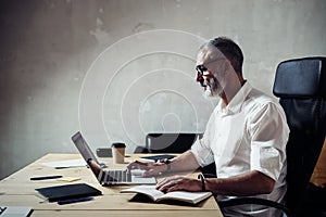 Adult bearded businessman wearing a classic glasses and working at the wood table in modern loft.Stylish middle age man