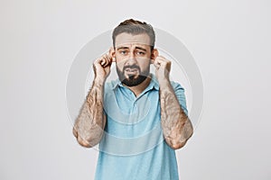 Adult bearded man with gloomy smile stretching ears, imitating sad monkey, over gray background. Guy is tired of hearing