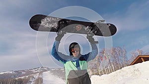 Adult bearded man standing in the ski park. He held up a snowboard and show that he was glad this day. He warmly dressed