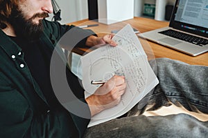 Adult bearded student sits with his feet up on his desk while studying online using laptop