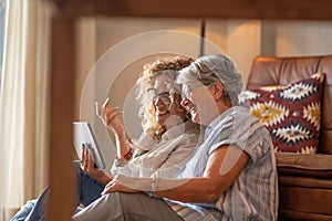 Adult beautiful daughter with elderly mother sitting at home looking social media on digital tablet