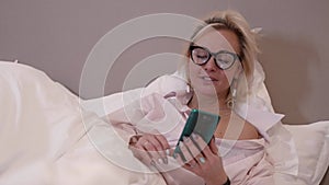 adult blonde woman with glasses is lying in bed before sleep and chatting by video call in cellphone