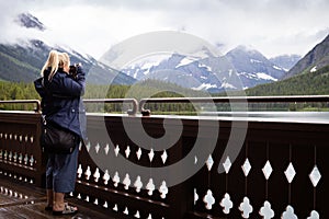 Adult blonde woman takes photos of Swiftcurrent Lake on the deck of the Many Glacier Hotel in Glacier National Park Montana USA
