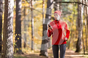 Adult breathless woman in red sportswear jogging outdoor with cellphone and wired headphones as a music player in the