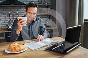 Adult businessman sit at table in kitchen anr read journal. He hold white cup and look down. Laptop and plate with