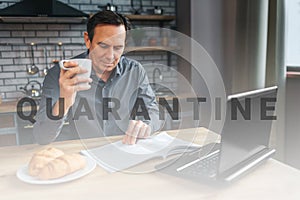 Adult businessman sit at table in kitchen anr read journal. He hold white cup and look down. Laptop and plate with