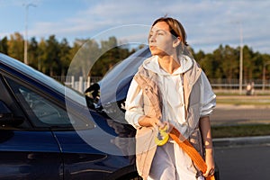 an adult Caucasian businesswoman with a cable for transporting a car stands at the raised hood of a broken car and waits